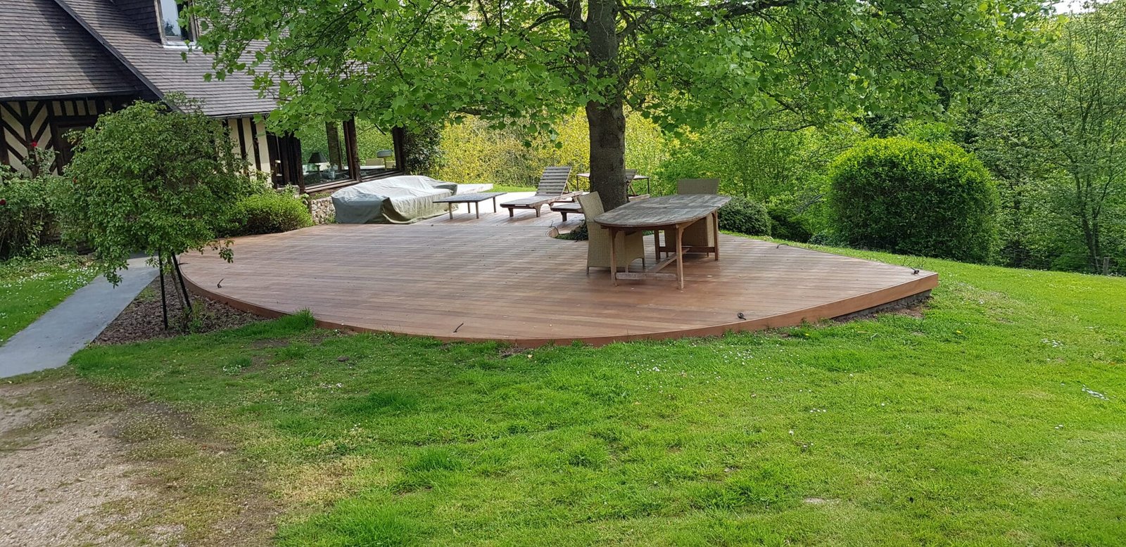 Vaste terrasse en bois en forme de demi-cercle avec mobilier de jardin, entourée de verdure et d'arbres, offrant une vue sur la nature.