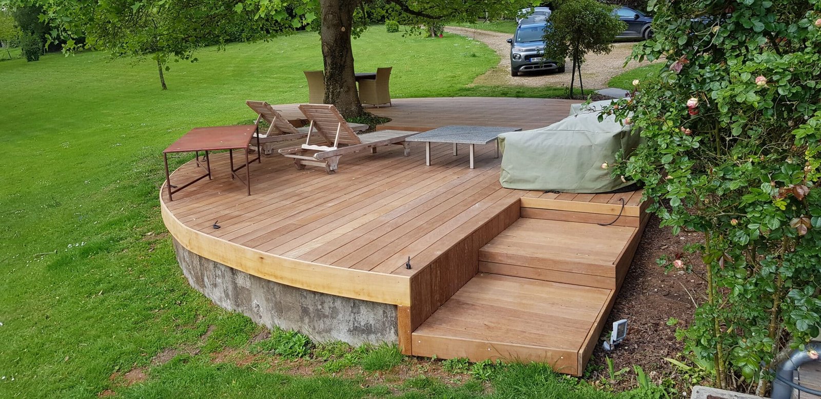 Terrasse en bois avec chaises longues et table basse dans un jardin verdoyant.