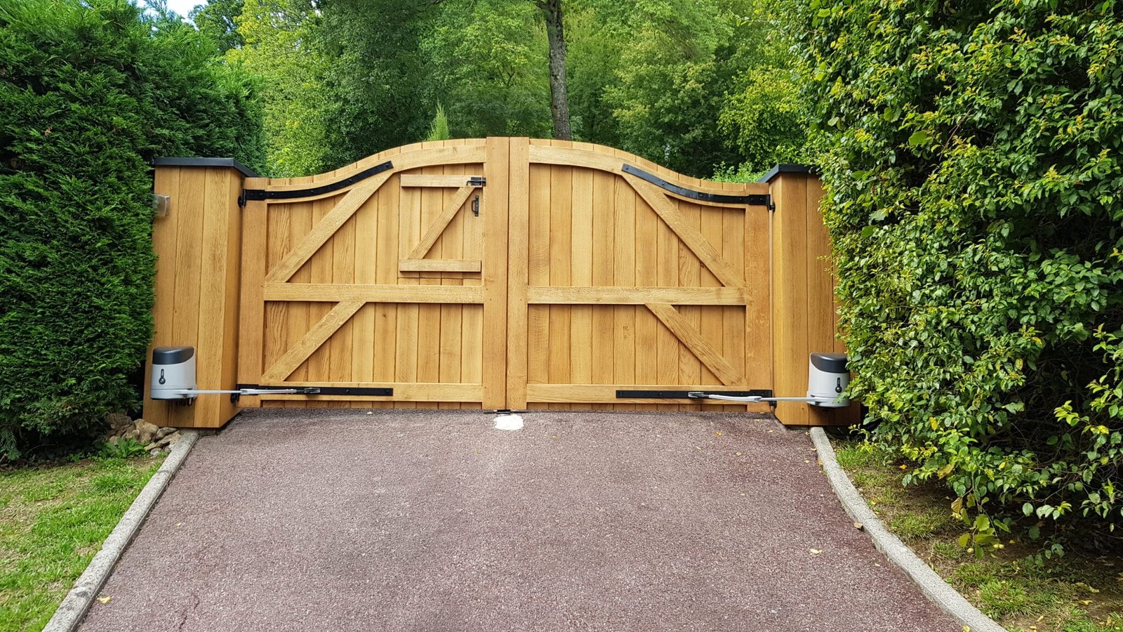 Grand portail en bois, encadré par des haies vertes, ouvrant sur une allée en béton.