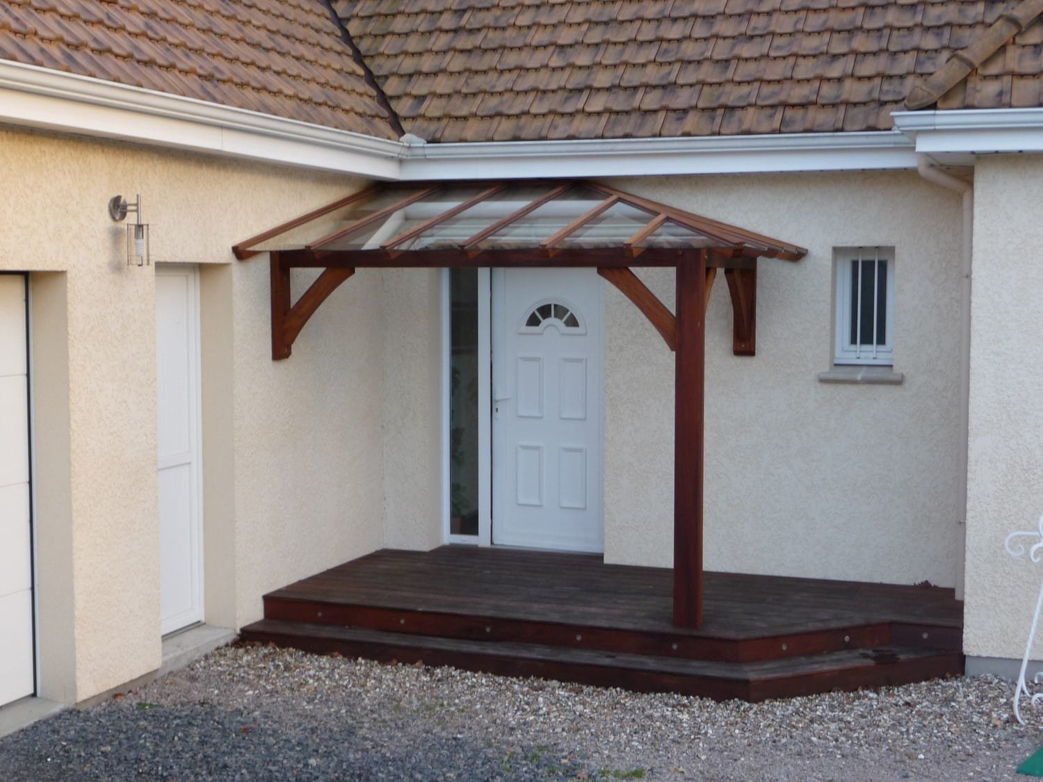 Entrée de maison avec une petite terrasse en bois, abritée par une marquise en bois et verre.