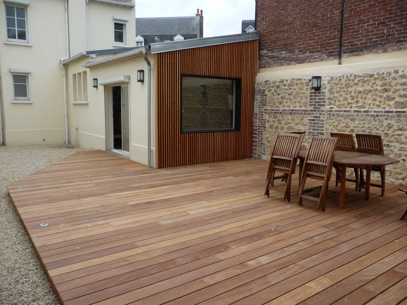 Terrasse en bois avec bardage vertical en bois et mur en pierre apparente, comprenant un ensemble de meubles de jardin en bois.