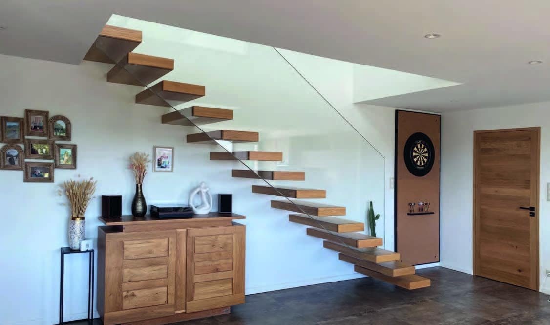 Un escalier flottant en bois avec une rampe en verre transparent dans un salon moderne. Le salon comprend un meuble en bois et des décorations murales. Le sol est en bois foncé.