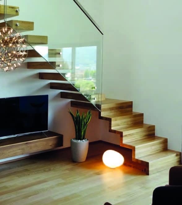 Un escalier flottant en bois clair avec une rampe en verre. L'escalier est situé dans un salon avec une télévision, une plante en pot et un sol en bois clair. Une lampe sphérique blanche est posée sur le sol.