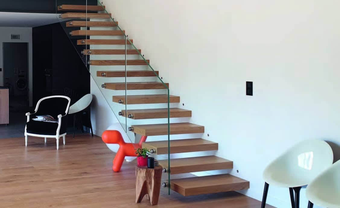 Un escalier flottant en bois avec une rampe en verre transparent. L'escalier se trouve dans un salon moderne avec des chaises blanches et une chaise noire. Le sol est en bois clair.