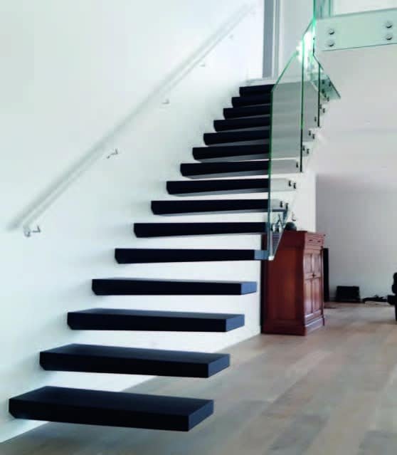 Un escalier flottant noir avec une rampe en verre dans un intérieur moderne. Le sol est en bois clair et les murs sont blancs. L'escalier mène à un niveau supérieur avec un mur blanc.