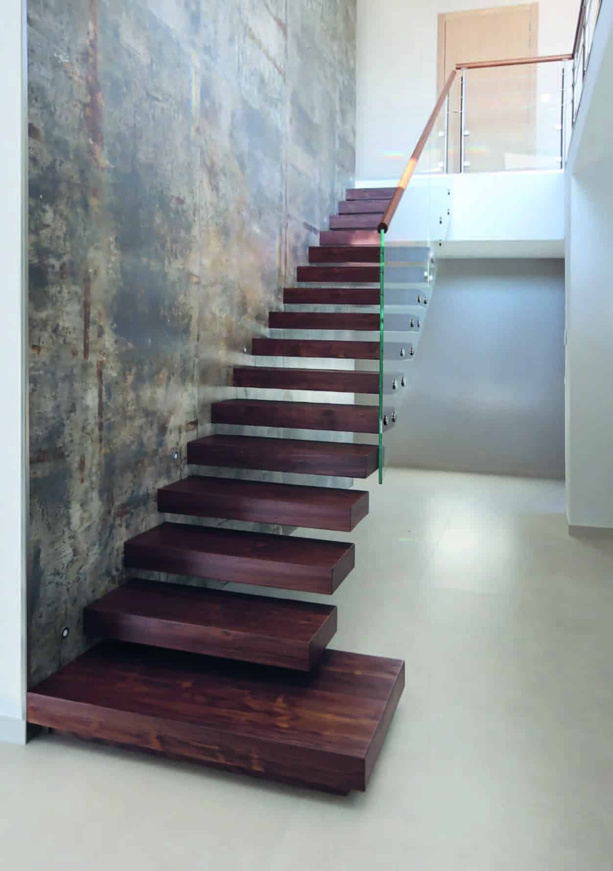 Un escalier flottant en bois foncé avec une rampe en verre. L'escalier se trouve dans un intérieur avec un mur texturé et un sol clair. L'escalier mène à un niveau supérieur avec une barrière en verre et bois.