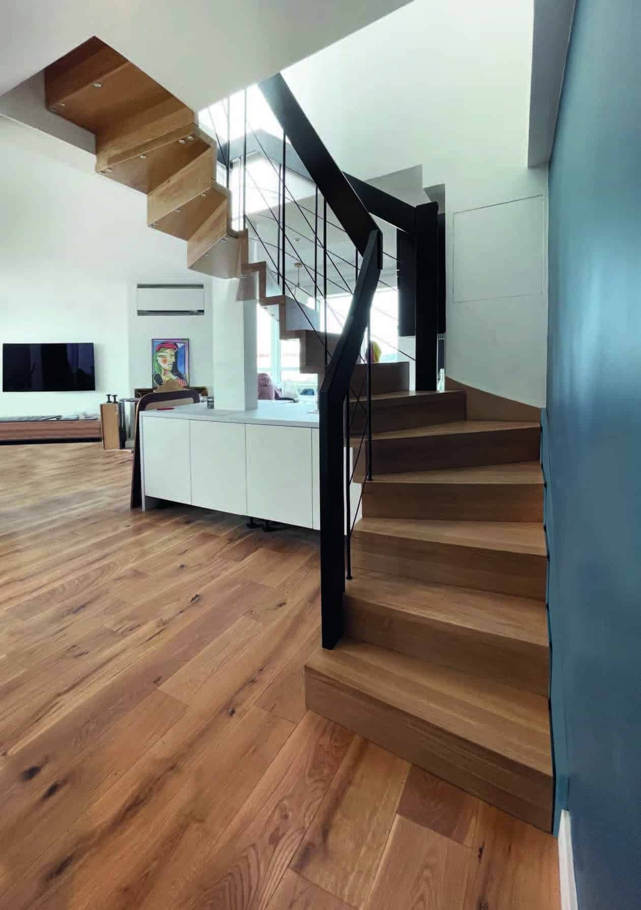 Escalier moderne avec des marches en bois et une rampe en métal noir, dans un espace de vie lumineux avec un parquet en bois.