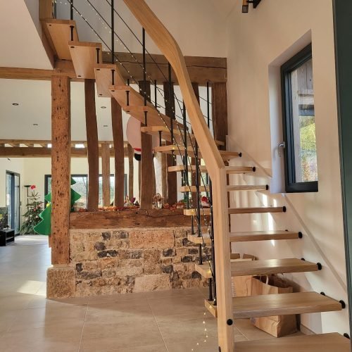 Escalier moderne avec des marches en bois et une structure en métal noir, accompagné d'une rampe en bois courbé, situé à côté d'un mur de pierres.