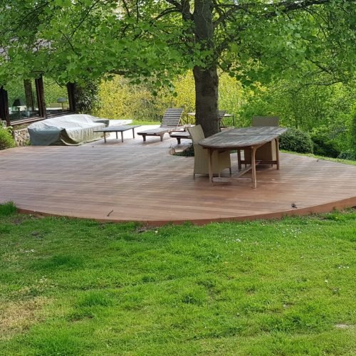 Vaste terrasse en bois en forme de demi-cercle avec mobilier de jardin, entourée de verdure et d'arbres, offrant une vue sur la nature.