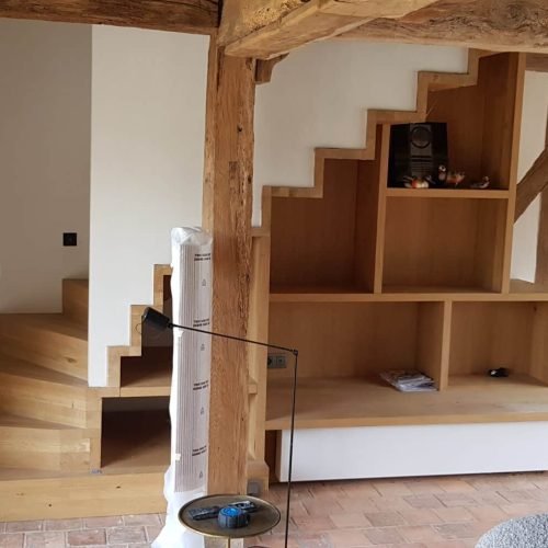 Un intérieur rénové présentant un escalier en bois intégré à des étagères et des espaces de rangement sur mesure, dans une maison avec des poutres apparentes.